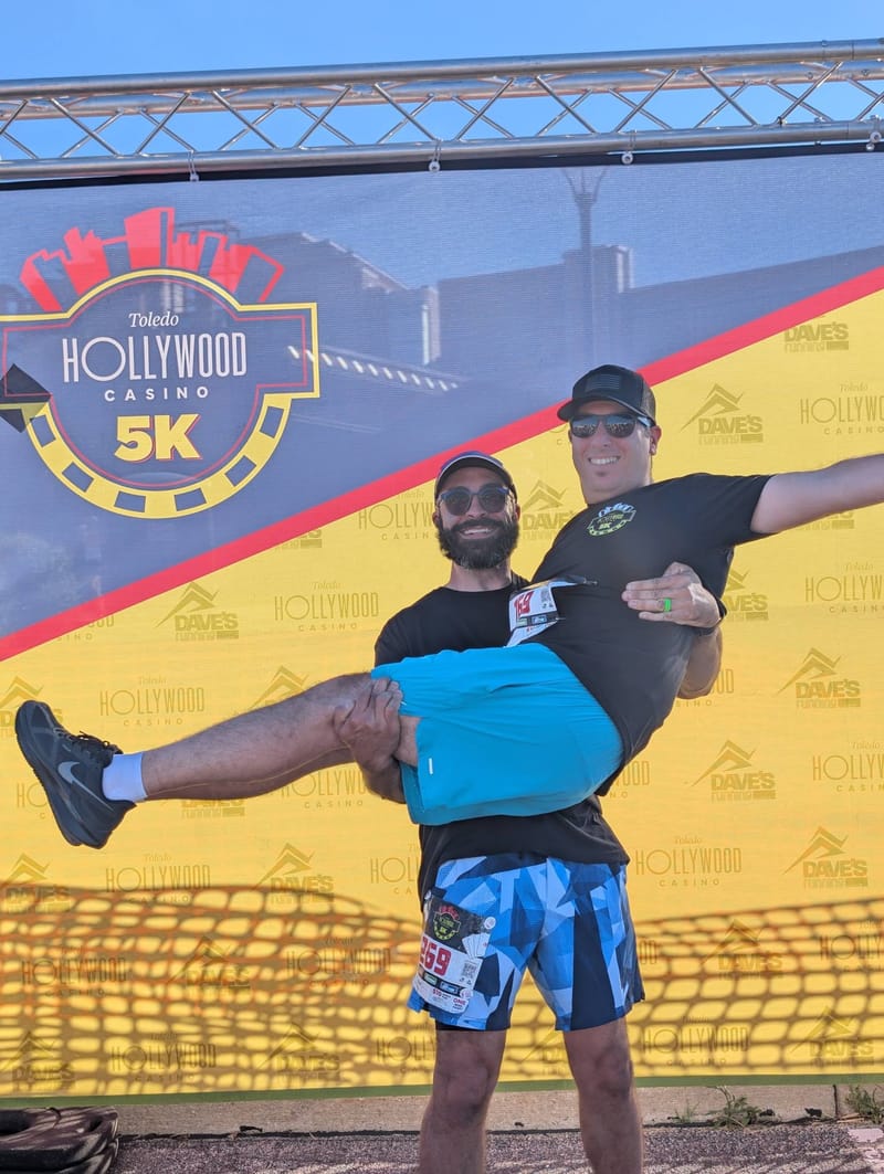 Abe being carried horizontally in another runner’s arms, both laughing, in front of a Toledo Hollywood Casino 5K backdrop.
