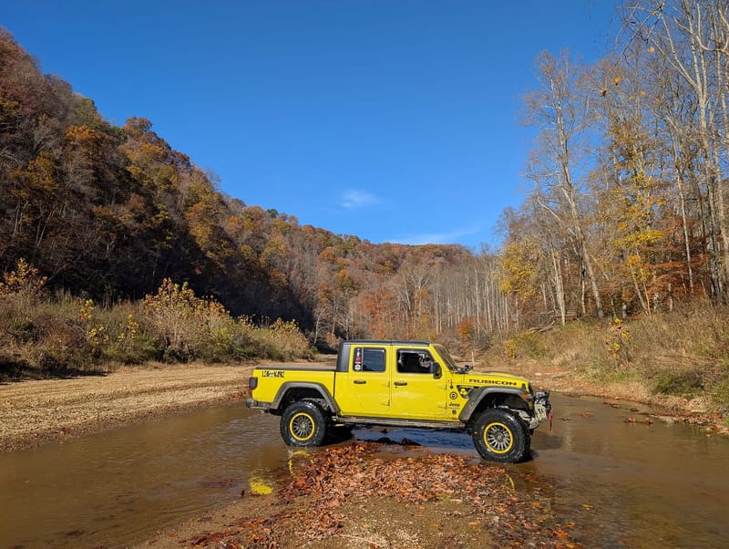 The yellow Jeep Gladiator standing in a shallow creek crossing, water around its tires, autumn hillside rising behind.