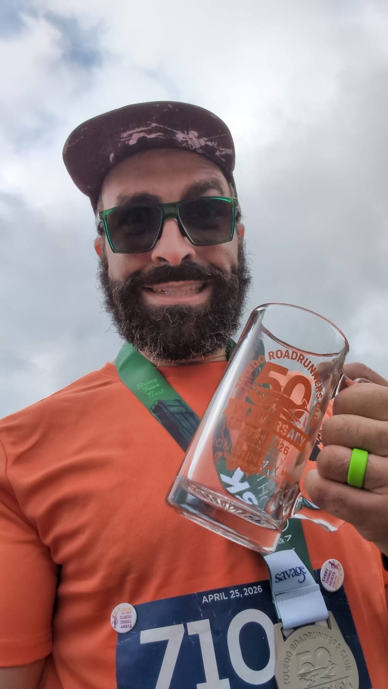 Post-race selfie under a gray sky, finisher medal around his neck and a commemorative glass mug held up, bib dated 25 April 2026.